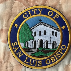 May include: A circular embroidered patch featuring the text CITY OF SAN LUIS OBISPO in yellow thread on a dark blue border. The centre depicts a white mission-style building with a cross, a green tree, and brown ground against a light blue sky.
