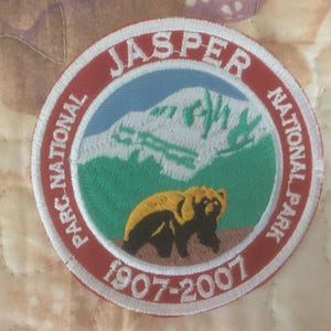 May include: Embroidered patch featuring a bear in front of a mountain scene. The patch is circular with the words "JASPER NATIONAL PARK" and "1907-2007" in white lettering on a red border. The bear is brown and black.