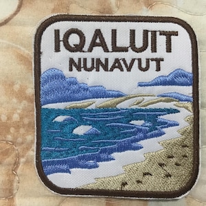 May include: A rectangular embroidered patch with rounded corners featuring a brown border. The text IQALUIT NUNAVUT is stitched in brown at the top. Below, a landscape depicts a blue ocean, a sandy beach, and light blue mountains.