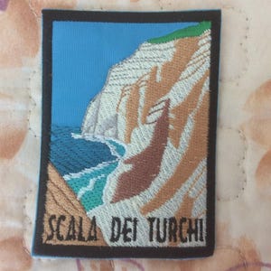 May include: Embroidered patch featuring a coastal landscape with a blue sky, turquoise water, and tan cliffs. The patch has a black border and the text "SCALA DEI TURCHI" in white.