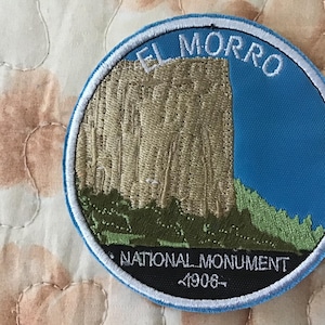 May include: A circular embroidered patch featuring a tan rock formation against a bright blue sky. The text EL MORRO is stitched in white at the top, with NATIONAL MONUMENT and 1906 stitched in white on a black background at the bottom.