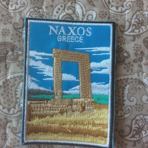 May include: Embroidered patch with the text "NAXOS GREECE" above a stone archway image. The patch has a blue background with white clouds, a tan arch, and a green and tan landscape.