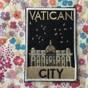 May include: Embroidered patch depicting the Vatican City skyline, with the words "VATICAN CITY" in gold and navy blue. The patch has a black background with white stars and a white border. It is set against a floral fabric.