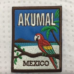 Op de afbeelding: Een rechthoekige geborduurde patch met een blauwe lucht, een wit zandstrand en een groene palmboom. Een rode, gele en groene papegaai zit op een bruine tak. De tekst AKUMAL is in het wit bovenaan geborduurd en MEXICO in het bruin onderaan.
