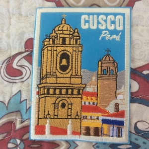 May include: Embroidered patch featuring a detailed illustration of a Cusco, Peru landmark. The design includes a tall, ornate building with a bell tower, a smaller tower, and the text "CUSCO Peru" against a blue sky background. The patch has a white border.