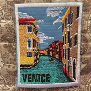 Op de afbeelding: Geborduurde patch met een kanaalscène in Venetië, Italië. De patch toont kleurrijke gebouwen, een waterweg en een gondel onder een blauwe lucht met wolken. Het woord "VENICE" is onderaan geborduurd.