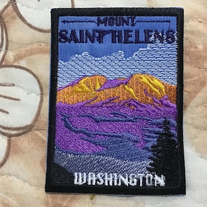 May include: A rectangular embroidered patch featuring a landscape of Mount Saint Helens. The design includes a blue sky, golden mountain peaks, purple slopes, and a black evergreen tree silhouette. The text reads MOUNT SAINT HELENS and WASHINGTON.