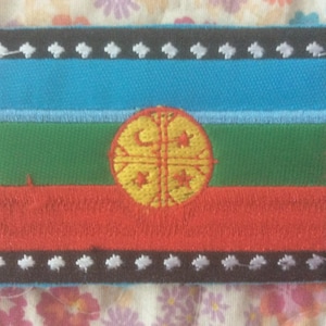 May include: Embroidered patch featuring a flag design with horizontal stripes in blue, green, and red. A yellow circular emblem with a cross-like pattern is centered on the green stripe. The patch has a black border with white star-shaped accents.