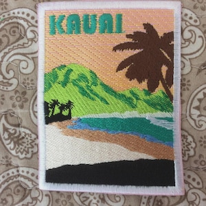 Pode incluir: Patch bordado com uma cena de praia tropical com a palavra "KAUAI" em verde. O design inclui uma montanha, palmeiras e uma praia com água azul e bege. O patch tem uma borda branca.