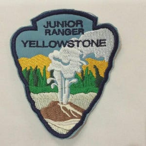 May include: Embroidered patch featuring a Yellowstone National Park design. The patch includes text that reads "JUNIOR RANGER YELLOWSTONE". The design depicts a geyser erupting, with trees, mountains, and a blue and yellow sky.