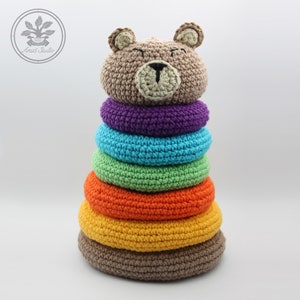 May include: A brown crocheted bear head sits atop a rainbow-colored stacking toy. The rings are purple, turquoise, green, orange, yellow, and brown.