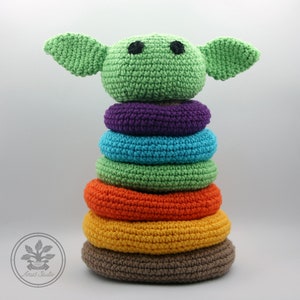 Może przedstawiać: Zabawka do układania w stos wykonana na szydełku w kształcie zielonego kosmity z dużymi uszami. Zabawka ma sześć kolorowych pierścieni w odcieniach fioletu, niebieskiego, zielonego, pomarańczowego, żółtego i brązowego.