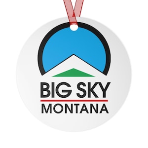 Op de afbeelding: Ronde witte ornament met een zwarte cirkel die een blauwe cirkel bevat met een witte bergtop en een groene basis. De tekst "BIG SKY MONTANA" is in zwart onder de cirkel gedrukt.