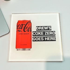 May include: A square white coaster featuring a red Coca-Cola Zero Sugar soda can graphic on the left side and black text on the right that reads DREW'S COKE ZERO GOES HERE in three stacked lines with black rectangular backgrounds behind each line.