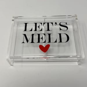 Puede incluir: Una bandeja de acrílico transparente con las palabras "LET'S MELD" en negro y un corazón rojo. La bandeja rectangular tiene tapa y está diseñada para almacenamiento o exhibición.