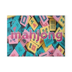 May include: A collection of rectangular mahjong tiles in shades of pink, yellow, and teal, each featuring unique black and red engraved symbols. The word mahjong is overlaid in large, bold, pink letters across the center of the tile arrangement.