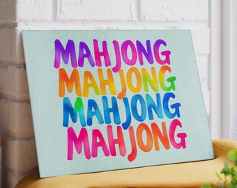 Puede incluir: Una tabla decorativa rectangular de color azul claro con la palabra "MAHJONG" repetida cuatro veces en un arcoíris de colores. La tabla está apoyada contra una pared de ladrillos blancos y una superficie amarilla.