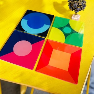 May include: Four colourful coasters with geometric designs. The coasters are square and feature a variety of colours, including blue, green, pink, orange, and yellow.