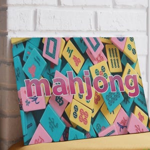 May include: A rectangular wall art print featuring a colorful collage of mahjong tiles in shades of pink, yellow, and teal. The word mahjong is printed in large, bold, magenta letters across the center of the tile pattern.