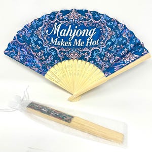 Könnte beinhalten: Ein Faltfächer mit blauem und rosa Blumenmuster und dem Text "Mahjong Makes Me Hot". Der Fächer hat einen Holzrahmen. Ein zweiter Fächer ist in einer durchsichtigen Tasche zu sehen.
