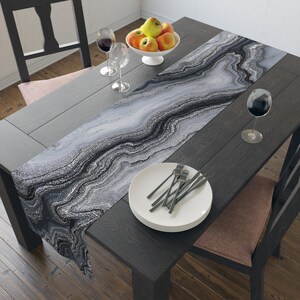 May include: A rectangular table runner featuring a black, white, and gray marbled agate pattern with swirling, wavy lines. The runner is laid out on a dark wooden table, accompanied by a stack of white plates, silverware, and wine glasses.