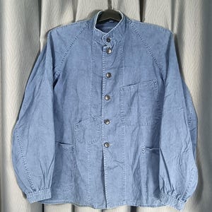 May include: A light blue denim jacket with a mandarin collar and button-up front. The jacket has long sleeves with gathered cuffs and multiple pockets. The garment is hanging on a black hanger against a neutral background.