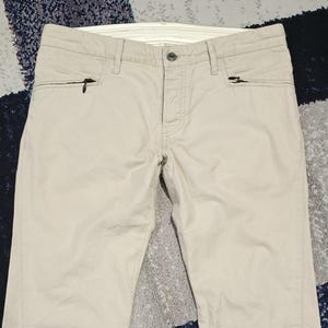 May include: Beige trousers with two zipped pockets on the front. The trousers have a button fastening and belt loops. The fabric looks to be a lightweight material, suitable for warmer weather. The trousers are neatly folded.