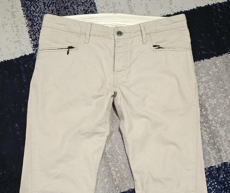 May include: Beige trousers with two zipped pockets on the front. The trousers have a button fastening and belt loops. The fabric looks to be a lightweight material, suitable for warmer weather. The trousers are neatly folded.
