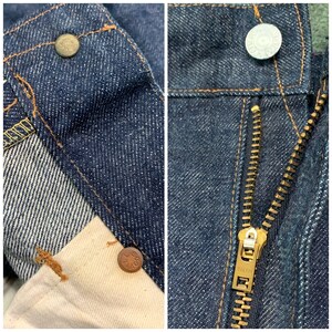 Pu&ograve; includere: Primo piano di un paio di jeans in denim blu con una cerniera in ottone e rivetti in rame. I jeans hanno uno stile classico a gamba dritta.