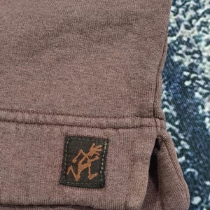 Pode incluir: Close-up de um moletom de cor malva. Um pequeno patch retangular marrom com uma figura estilizada &eacute; costurado na bainha inferior. O tecido tem uma apar&ecirc;ncia texturizada.