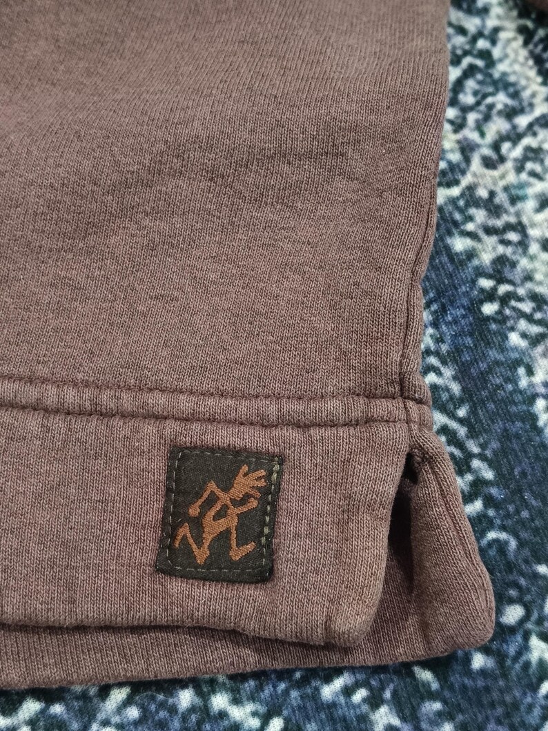 Pode incluir: Close-up de um moletom de cor malva. Um pequeno patch retangular marrom com uma figura estilizada &eacute; costurado na bainha inferior. O tecido tem uma apar&ecirc;ncia texturizada.