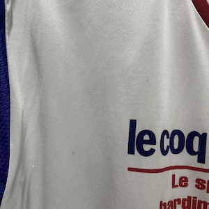 May include: White athletic jersey with blue and red trim. The jersey features the text "le coq" in navy blue, with additional red text below. The fabric has a subtle texture.