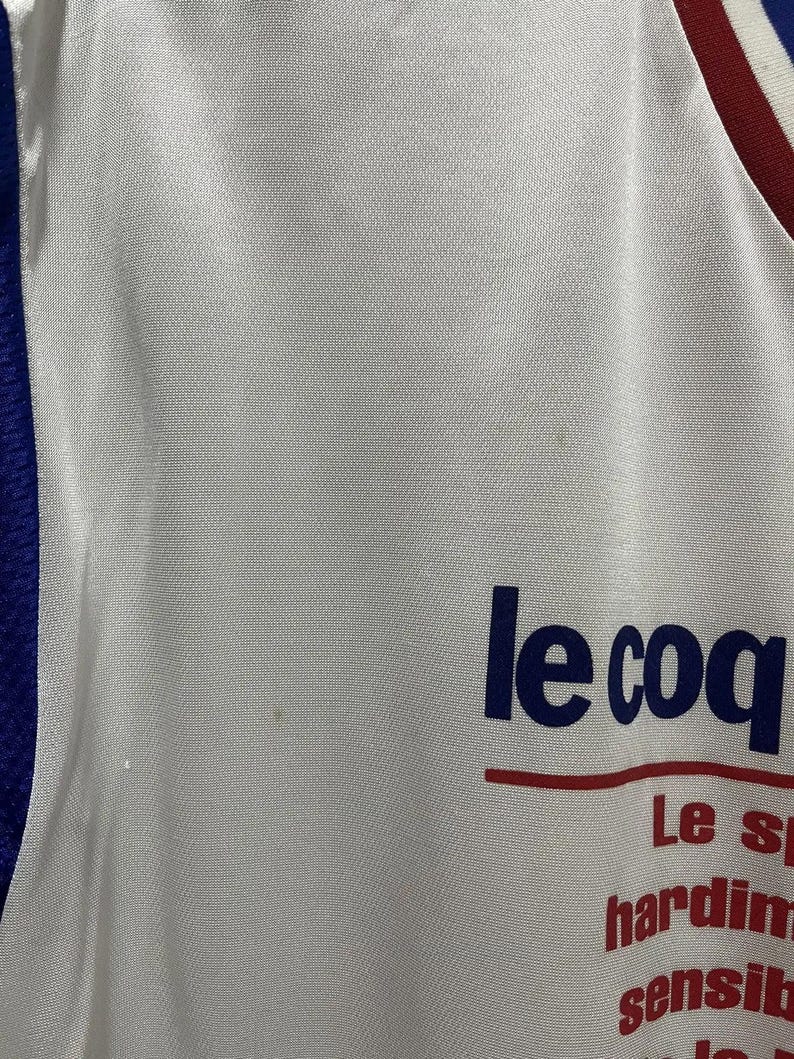 May include: White athletic jersey with blue and red trim. The jersey features the text "le coq" in navy blue, with additional red text below. The fabric has a subtle texture.