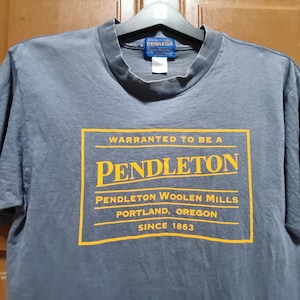 May include: A gray short-sleeved t-shirt with a yellow rectangular graphic on the front. The graphic reads "Warranted to be a Pendleton" with additional text indicating the company's name, location, and founding year, 1863.