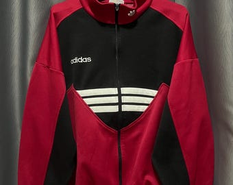 Könnte beinhalten: Rote, schwarze und weiße Adidas-Trainingsjacke mit durchgehendem Reißverschluss. Die Jacke hat das Adidas-Logo auf der Brust und drei weiße Streifen auf der Vorderseite. Die Jacke hängt an einem Kleiderbügel.