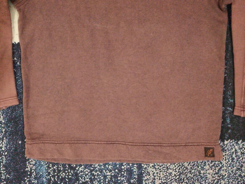 Pode incluir: Uma camisa marrom de manga comprida e gola redonda. A camisa tem uma apar&ecirc;ncia texturizada e um pequeno patch com o logotipo perto da bainha. As mangas s&atilde;o de um tom marrom ligeiramente mais claro do que o corpo.