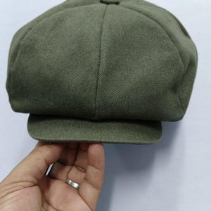 Könnte beinhalten: Eine olivgrüne Newsboy-Cap mit einer abgerundeten Krone und einem kurzen, steifen Schirm. Die Kappe besteht aus einem strukturierten Stoff. Die Farbe der Kappe ist durchgehend. Die Kappe wird von einer Hand gehalten.