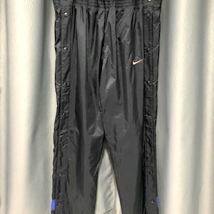 May include: Black nylon track pants with snap closures down the sides. The pants have an elastic waistband and a small orange Nike logo. Blue accents are visible near the ankles. The pants are hanging on a metal hanger.