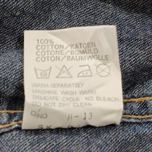 Pode incluir: Close-up de uma etiqueta de roupa branca com texto preto sobre um fundo de ganga. A etiqueta diz "100% COTTON" e inclui instru&ccedil;&otilde;es de lavagem. A etiqueta tamb&eacute;m tem o texto "H-13".