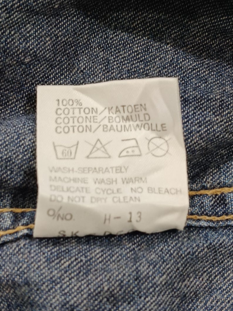 Pode incluir: Close-up de uma etiqueta de roupa branca com texto preto sobre um fundo de ganga. A etiqueta diz "100% COTTON" e inclui instru&ccedil;&otilde;es de lavagem. A etiqueta tamb&eacute;m tem o texto "H-13".