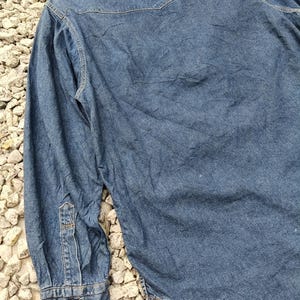 Pode incluir: Camisa de ganga azul m&eacute;dia com mangas compridas. A camisa apresenta uma frente abotoada, uma gola pontiaguda e punhos abotoados. A parte de tr&aacute;s da camisa tem um detalhe de pala. O tecido tem um aspeto ligeiramente gasto.
