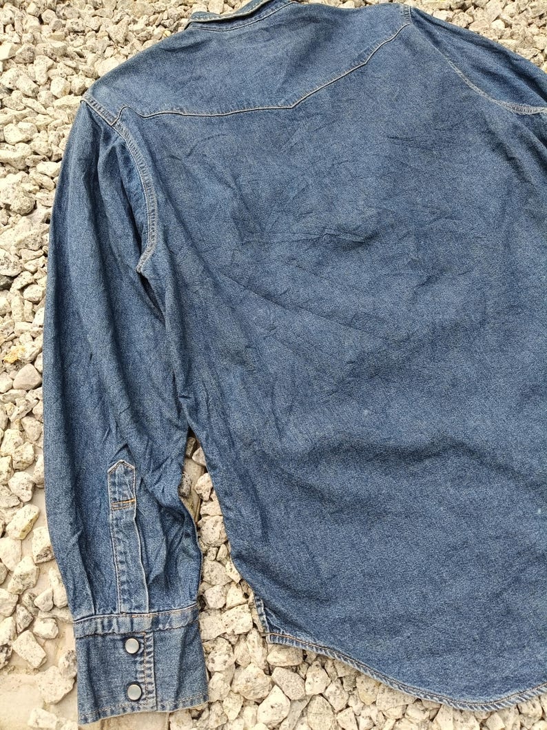 Pode incluir: Camisa de ganga azul m&eacute;dia com mangas compridas. A camisa apresenta uma frente abotoada, uma gola pontiaguda e punhos abotoados. A parte de tr&aacute;s da camisa tem um detalhe de pala. O tecido tem um aspeto ligeiramente gasto.