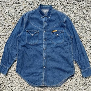 Pode incluir: Uma camisa de ganga de manga comprida em lavagem azul m&eacute;dio. A camisa apresenta dois bolsos no peito com abas pontiagudas, uma frente abotoada e uma gola cl&aacute;ssica. Uma pequena etiqueta amarela &eacute; vis&iacute;vel num dos bolsos.