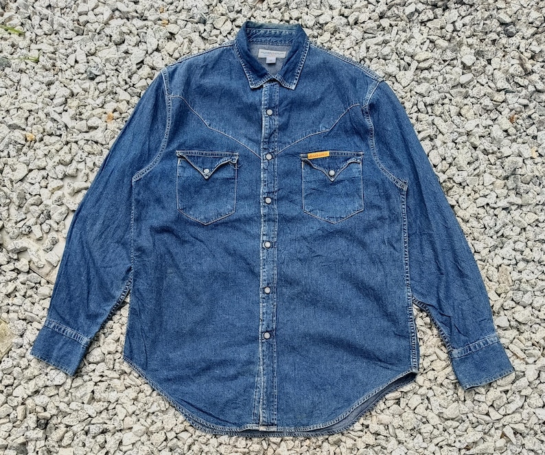 Pode incluir: Uma camisa de ganga de manga comprida em lavagem azul m&eacute;dio. A camisa apresenta dois bolsos no peito com abas pontiagudas, uma frente abotoada e uma gola cl&aacute;ssica. Uma pequena etiqueta amarela &eacute; vis&iacute;vel num dos bolsos.