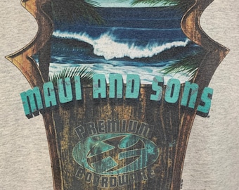 Può includere: T-shirt grigia con una grafica di uno scudo di legno con una scena di spiaggia. Lo scudo ha le parole "MAUI AND SONS" in lettere turchesi sopra le parole "PREMIUM BOARDWARE". L'immagine raffigura un oceano blu con onde e foglie di palma.