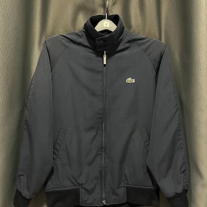 Puede incluir: Chaqueta azul marino con cuello, puños y dobladillo negros. La chaqueta tiene cierre de cremallera completo y un pequeño logotipo de cocodrilo verde en el pecho. La chaqueta está colgada de una percha.