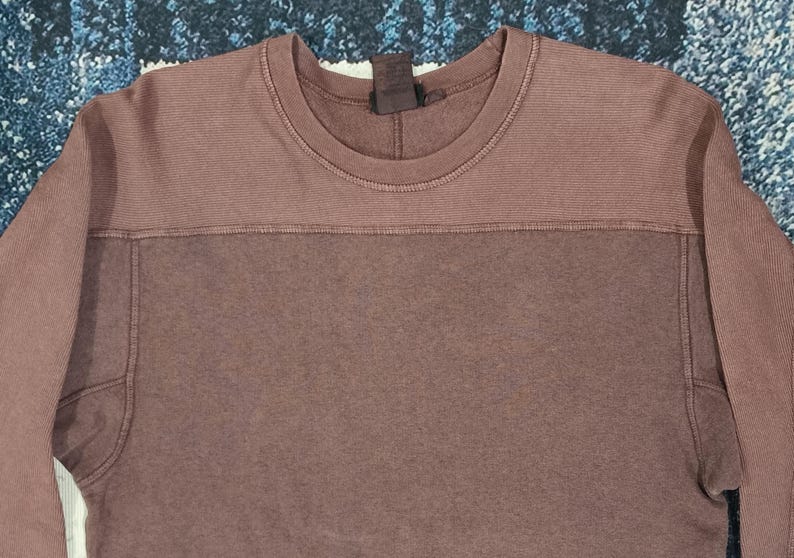 Pode incluir: Uma camisola de cor malva com gola redonda e mangas compridas. A sec&ccedil;&atilde;o superior da camisa tem uma textura canelada, enquanto a sec&ccedil;&atilde;o inferior &eacute; lisa. A camisa tem uma etiqueta na parte de tr&aacute;s do pesco&ccedil;o.