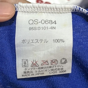 May include: A white tag with the text "QS-0684", "95S 101-4N", and "ポリエステル 100%". The tag also includes care symbols and Japanese text. The tag is attached to a blue fabric.