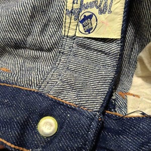 May include: Close-up of denim fabric with a visible label. The label reads "Wrangler CANADA SANFORIZED 12" with a blue and white logo. The jeans have a button with the number "12" on it and orange stitching.