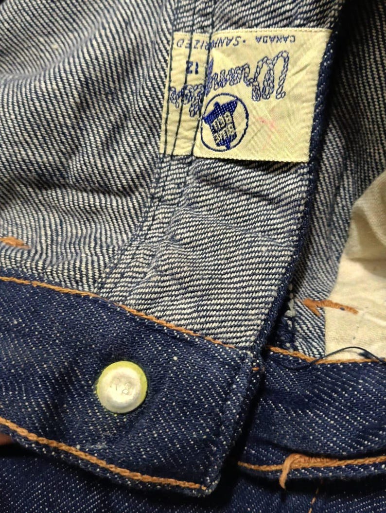May include: Close-up of denim fabric with a visible label. The label reads "Wrangler CANADA SANFORIZED 12" with a blue and white logo. The jeans have a button with the number "12" on it and orange stitching.